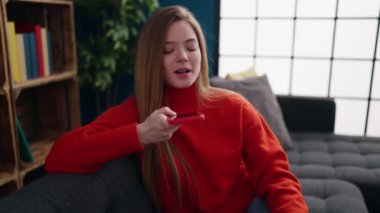 Genç sarışın kadın akıllı telefonda konuşuyor. Evdeki kanepede oturuyor.