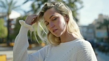 Genç sarışın kadın ciddi bir ifadeyle parkta saçlarına dokunuyor.