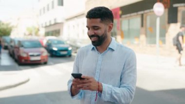 Genç Arap adam sokakta akıllı telefon kullanarak kendinden emin gülümsüyor.