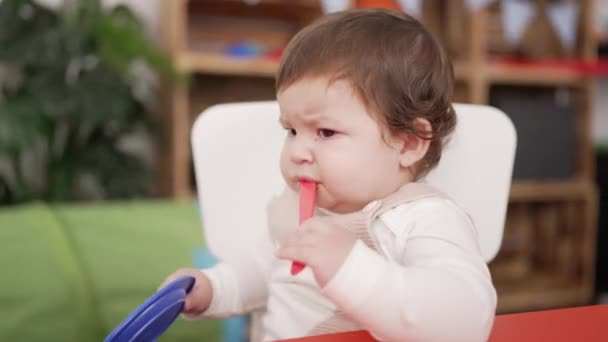 可爱的幼儿坐在桌子上吸吮着塑料勺子 手里拿着幼稚园里的盘子 — 图库视频影像
