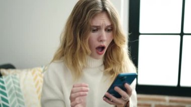 Genç sarışın kadın evde akıllı telefon kullanıyor.