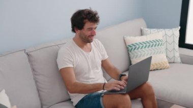 Genç İspanyol adam evdeki koltukta dizüstü bilgisayar kullanıyor.