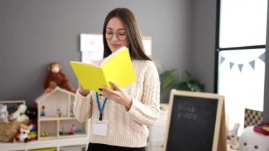 Young beautiful hispanic woman preschool teacher reading book standing at kindergarten