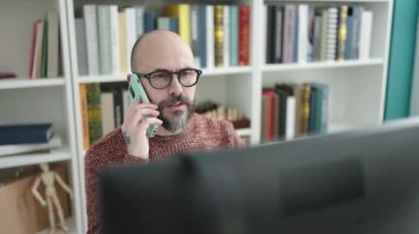 Genç, kel bir öğrenci üniversite sınıfında bilgisayar kullanarak akıllı telefondan konuşuyor.