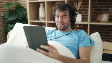 Young hispanic man using touchpad and headphones lying on bed at bedroom