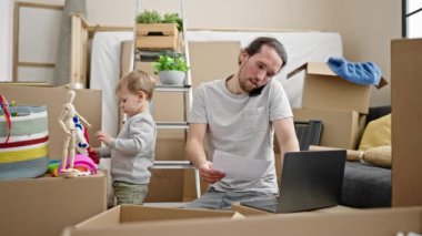 Baba ve oğul yeni evlerinde dizüstü bilgisayarlarını kullanarak karton kutuları açıyor.