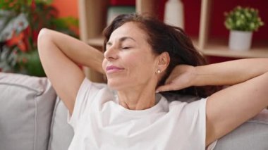 Orta yaşlı İspanyol kadın, elleri başının üstünde, evinde kanepede oturuyor.