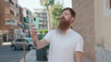 Genç kızıl saçlı adam gülümsüyor. Kendine güveni tam. Sokakta akıllı telefonun yanında selfie çekiyor.