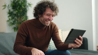 Young hispanic man watching video on touchpad sitting on sofa at home