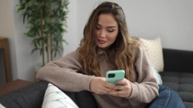 Young beautiful hispanic woman using smartphone doing ok gesture at home