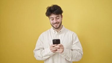 Zeki telefon kullanan genç İspanyol adam izole edilmiş sarı arka planda gülümsüyor.