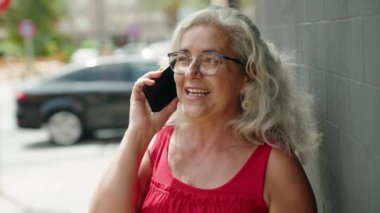 Orta yaşlı, kır saçlı, gülümseyen, kendine güvenen bir kadın caddedeki akıllı telefondan konuşuyor.