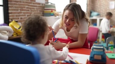 Teacher and toddler sitting on table drawing on paper at kindergarten