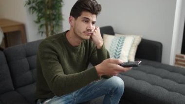 Young hispanic man watching tv sitting on sofa with boring expression at home