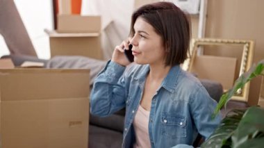 Genç beyaz kadın kanepede oturuyor ve yeni evinde telefonla konuşuyor.