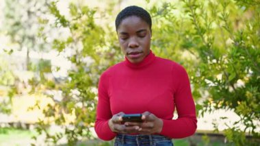 Parkta gülümseyen akıllı telefon kullanan Afrikalı Amerikalı kadın.
