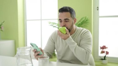Young hispanic man eating apple using smartphone at home