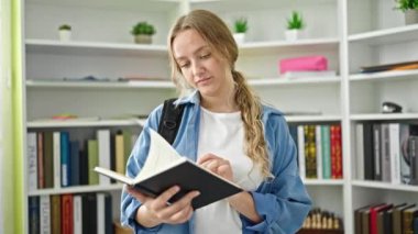 Young blonde woman student reading book doing silence gesture at library university
