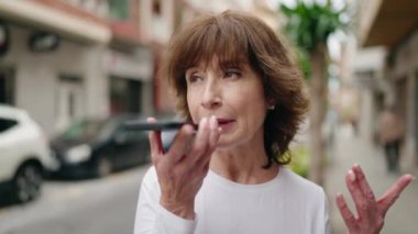 Orta yaşlı bir kadın gülümsüyor. Sokakta akıllı telefondan konuşuyor.