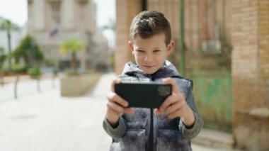 Sarışın çocuk sokakta akıllı telefonun yanında video oyunu oynuyor.