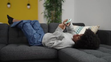 Afro-Amerikalı kadın akıllı telefon kullanıyor. Evdeki koltukta yatıyor.