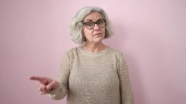 Gri saçlı orta yaşlı bir kadın, izole edilmiş pembe arka planda parmağıyla hayır diyor.
