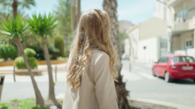 Genç sarışın kadın gülümsüyor. Parkta kollarını kavuşturmuş duruyor.