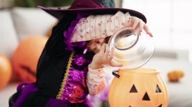 Adorable hispanic girl having halloween party putting sweets on pumpkin basket at home