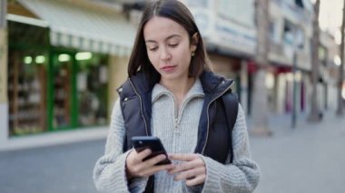 Genç, güzel, İspanyol bir kadın sokakta akıllı telefon kullanarak gülümsüyor.