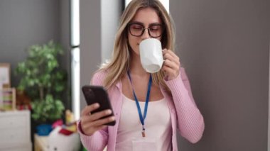 Young blonde woman preschool teacher using smartphone drinking coffee at kindergarten