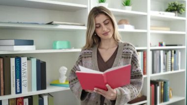 Young blonde woman teacher reading book doing silence gesture at library university