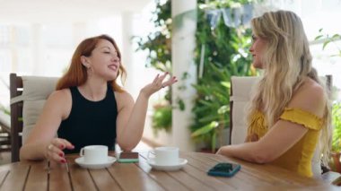 Akıllı telefon kullanan ve kahve içen iki kadın evdeki terasta oturuyor.