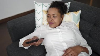 African american woman watching tv lying on sofa with boring expression at home