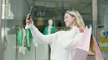 Sarışın genç bir kadın alışveriş yapmaya gidiyor. Elinde çantalar var. Sokakta akıllı telefonlardan selfie çekiyor.
