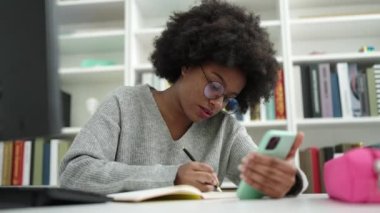 African american woman student using smartphone writing on notebook at library university