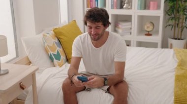 Young hispanic man using smartphone sitting on bed at bedroom