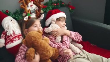 Erkek ve kız kardeş oyuncak ayılara sarılıyor. Evde Noel süslemeleriyle koltukta oturuyorlar.