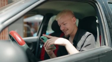 Beyaz bir adam akıllı telefon kullanıyor. Sokakta arabada oturuyor.