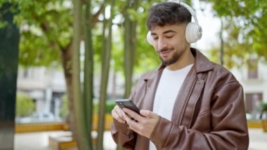 Genç Arap adam kendinden emin bir şekilde gülümsüyor. Parkta müzik dinliyor ve dans ediyor.