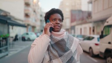 Genç Afrikalı Amerikalı kadın gülümsüyor. Kendine güveni tam. Sokakta akıllı telefondan konuşuyor.