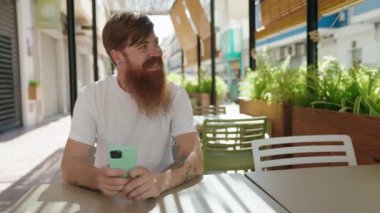 Genç kızıl saçlı adam kahve dükkanının terasında oturan akıllı telefonu kullanıyor.