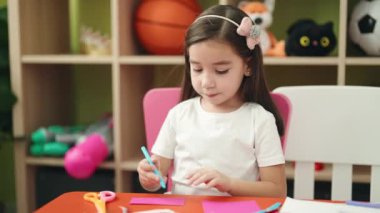 Adorable hispanic girl preschool student sitting on table drawing on paper at kindergarten