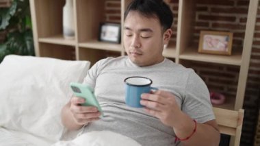 Genç Çinli adam akıllı telefon kullanıyor. Yatak odasında kahve içiyor.