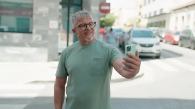 Orta yaşlı, gri saçlı, gülümseyen, kendine güvenen, akıllı telefondan selfie çeken bir adam.