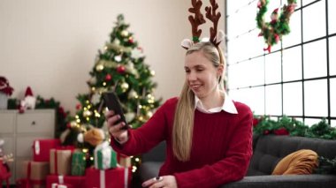 Genç sarışın kadın akıllı telefonuyla selfie çekiyor evdeki noel ağacının yanında koltukta oturuyor.