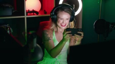 Young beautiful hispanic woman streamer playing video game using smartphone at gaming room
