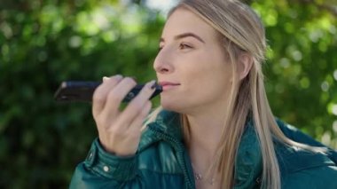 Young blonde woman sending voice message with smartphone at park