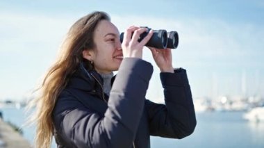 Genç beyaz kadın deniz kenarında dürbün kullanıyor.