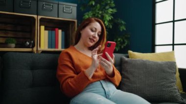 Genç kızıl saçlı neşeli kadın akıllı telefon kullanarak evde kutlama yapıyor.