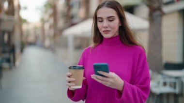 Genç sarışın kadın akıllı telefon kullanıyor. Sokakta kahve içiyor.